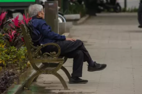 Starší muž sedí sám na lavičce v parku a zamyšleně se dívá do dálky