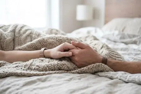 Jemná a detailní fotografie, na které se propletené ruce muže a ženy, částečně schované pod pletenou dekou, hřejí o sebe, a která je laděna do světlých, neutrálních tónů.