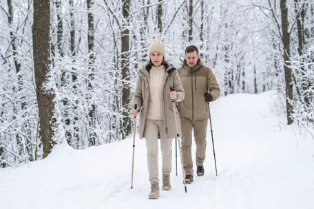 Dvojice lidí při nordic walkingu se sportovními holemi na zasněžené lesní cestě.